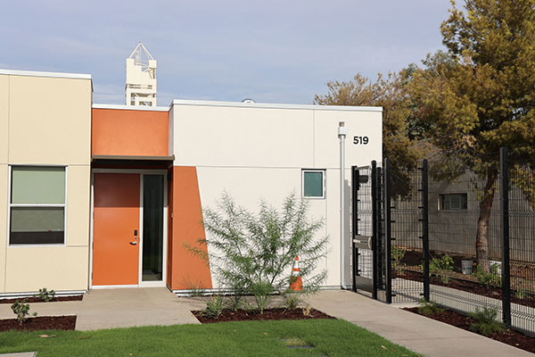 Exterior of a modern campus building with an orange entrance door, landscaped lawn, trees, and a fenced outdoor area.