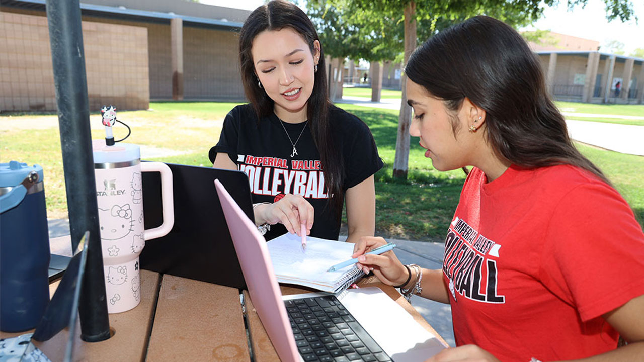 Two Imperial Valley College students study together outdoors, reviewing notes beside laptops at a campus table