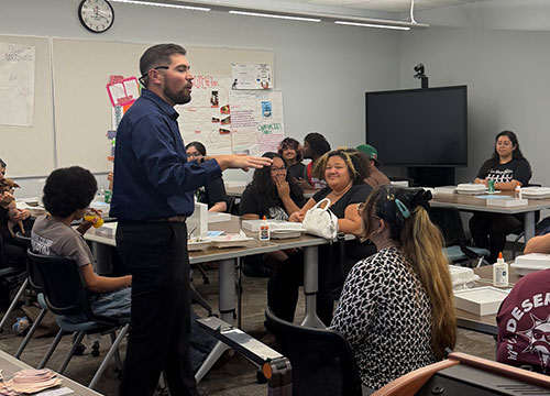 A presenter stands and speaks to a group of seated students around tables in a classroom setting.