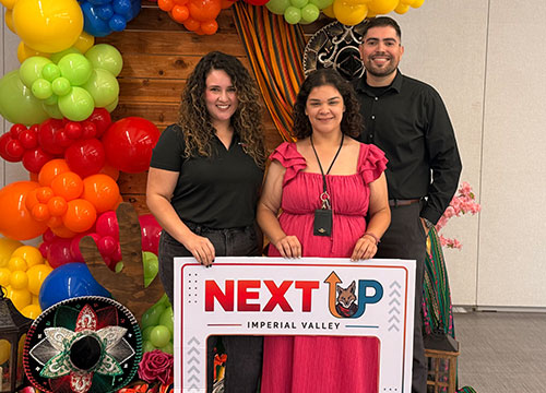Imperial Valley College NextUp staff Alexa Villagrana, Lesly Tirado, and Eduardo Pesqueira posing together behind a "NextUp Imperial Valley" event frame, in front of a colorful balloon arch and festive decorations.