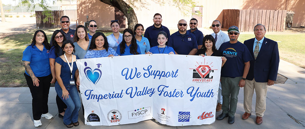 Group portrait of Imperial Valley College faculty, staff, and leadership standing outdoors on campus holding a large white banner that reads "We Support Imperial Valley Foster Youth," featuring various department logos and the IVC 60th Anniversary emblem.